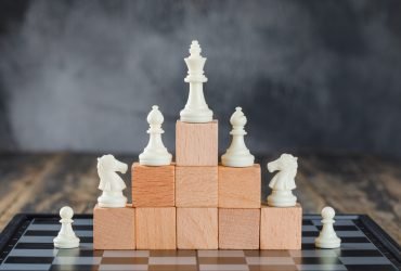 Business hierarchy concept with chessboard, figures on pyramid of wooden blocks on foggy and wooden background side view.