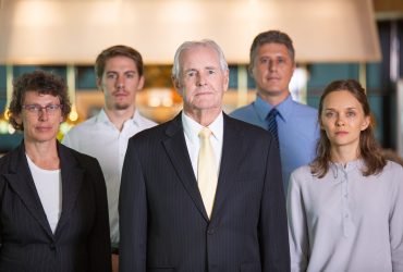 Closeup of business team of five serious diverse people standing and looking at camera with blurred view in background. Two blurred businessmen are in background. Front view.
